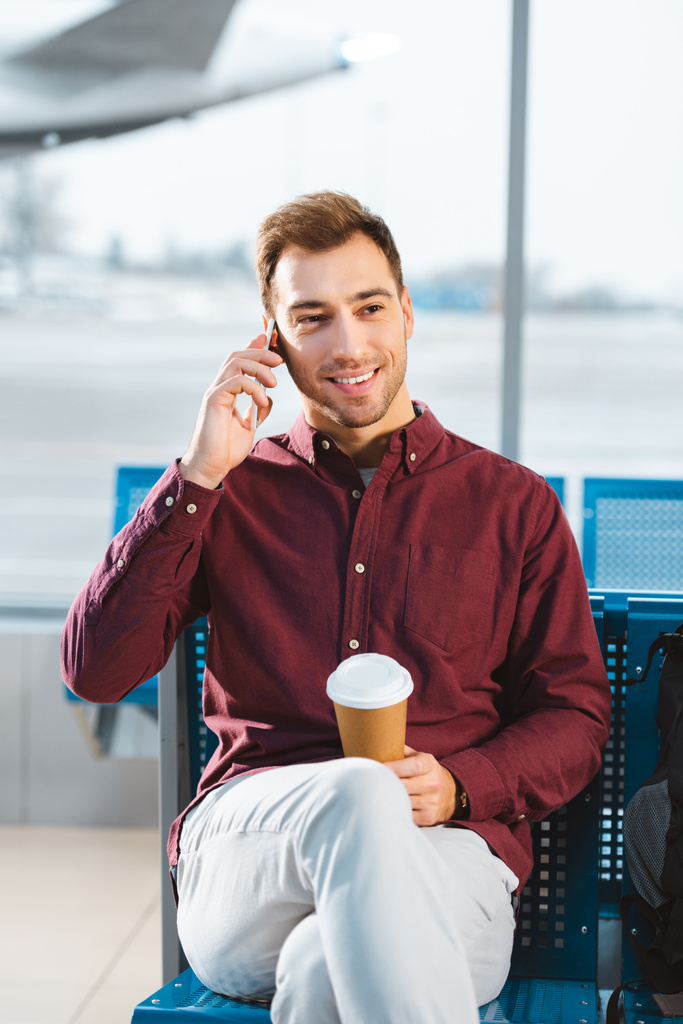 красивый мужчина разговаривает по смартфону и держит бумажную чашку в руках в аэропорту
  - Фото, изображение