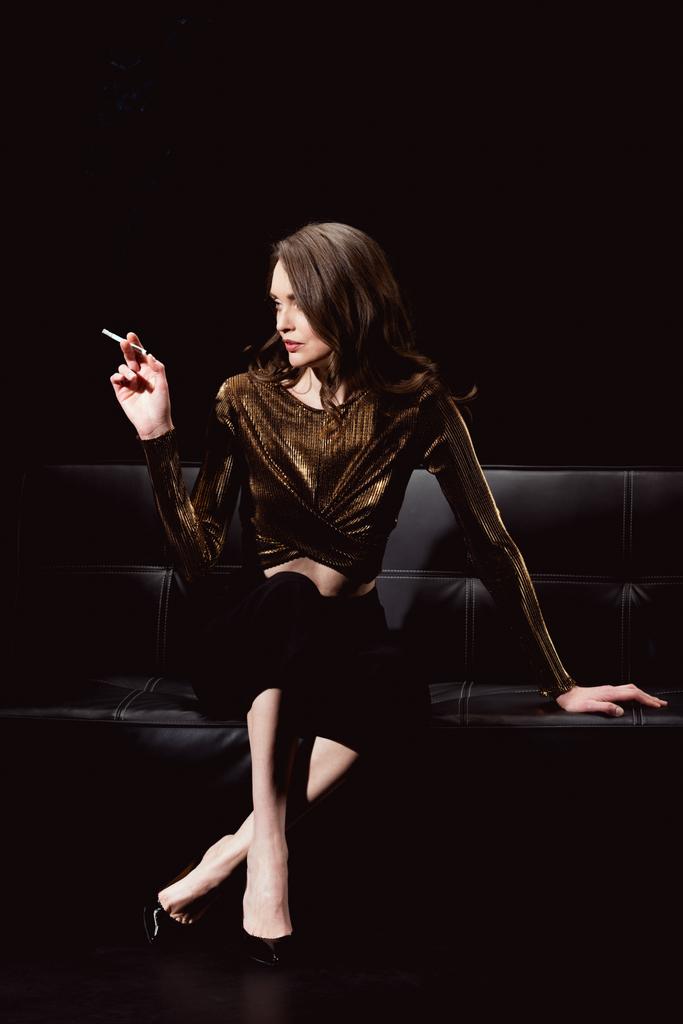 glamorous woman sitting on couch and smoking cigarette isolated on black - Photo, Image