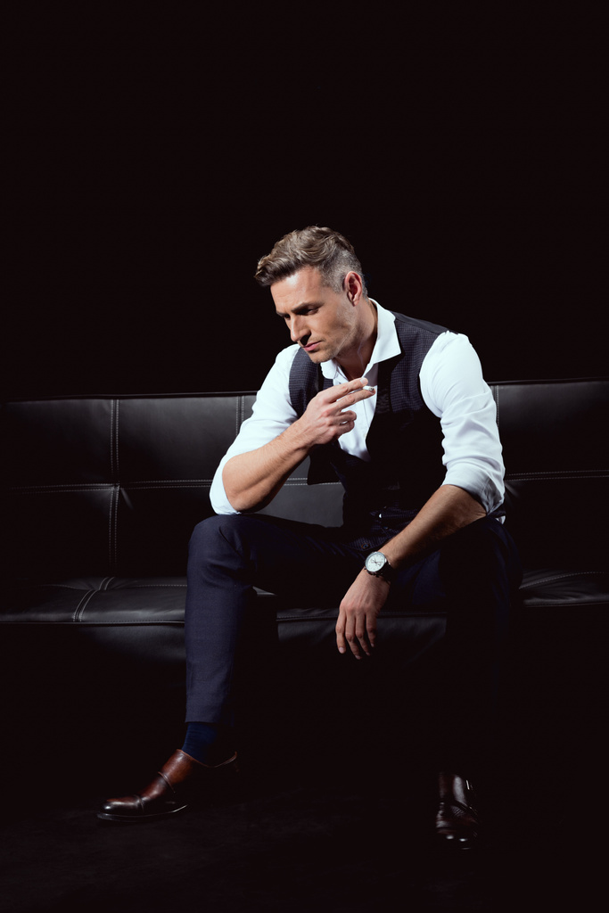hombre guapo serio en ropa formal sentado en el sofá con el cigarrillo aislado en negro
 - Foto, imagen