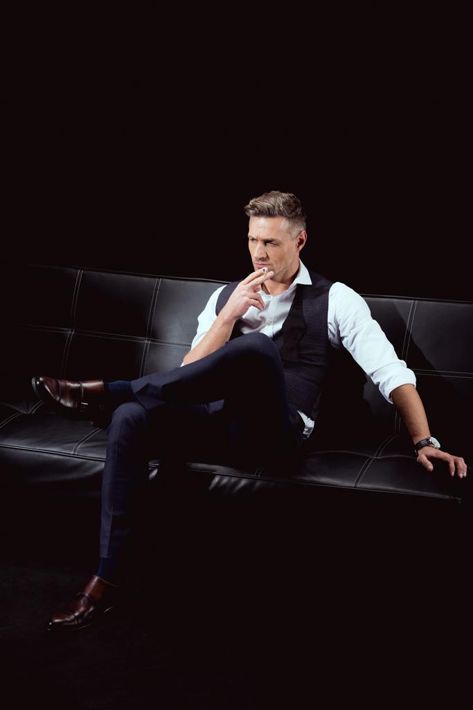 serious handsome man in formal wear sitting on couch with cigarette isolated on black - Photo, Image