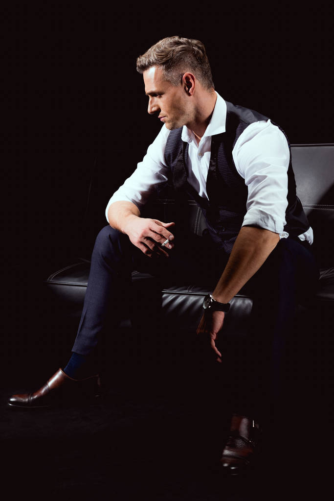 Красивый мужчина в формальной одежде сидит на диване с сигаретой, изолированной на черном
 - Фото, изображение