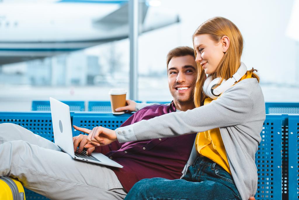 cheerful woman holding paper cup while pointing with finger at laptop near boyfriend in airport - Photo, Image