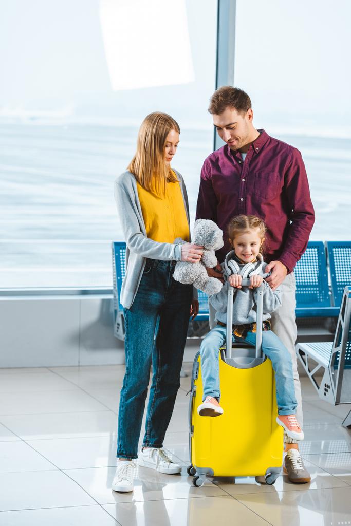 adorable child sitting on suitcase while mother holding teddy bear and standing near husband in waiting hall  - Photo, Image