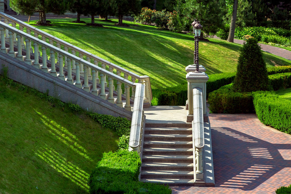 Uma escadaria de pedra com balaustradas railing para baixo no jardim com gramados verdes paisagísticos e arbustos cortados
. - Foto, Imagem