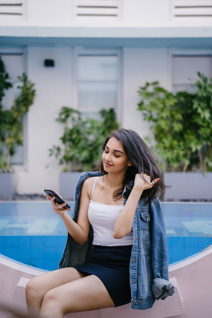 Een lifestyle portret van een jonge Indiase Aziatische vrouw zittend door een resort zwembad tijdens de dag terwijl het bedrijf smartphone - Foto, afbeelding