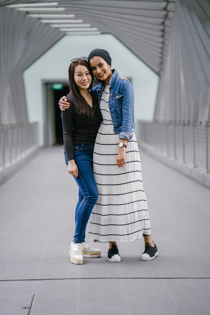 Двоє друзів різного етнічного походження на мосту протягом дня. Одна з них - малайська мусульманка, інша - китайська азіатка. Вони молоді, енергійні та привабливі.. - Фото, зображення