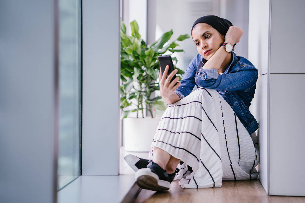 Egy fiatal közel-keleti muszlim nő ül a sarokban egy iroda, coworking tér vagy könyvtár a nap folyamán, és használja az okostelefonját. Fejkendőt visel (hidzsáb). - Fotó, kép
