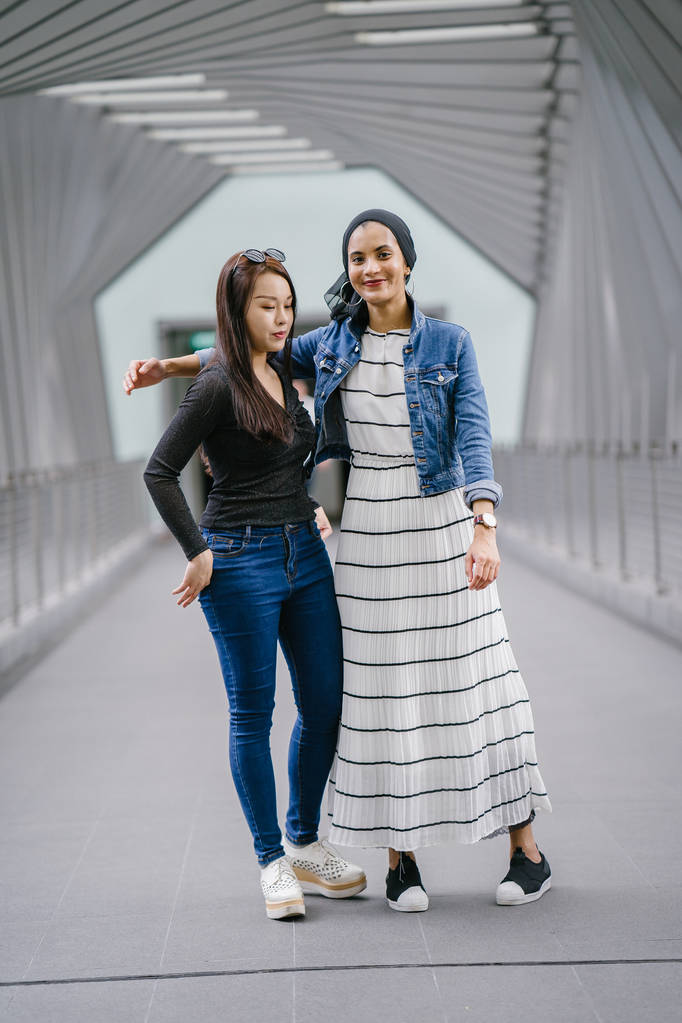 Zwei Freunde unterschiedlicher ethnischer Zugehörigkeit tagsüber auf einer Brücke. Die eine ist malaiische Muslimin, die andere asiatische Chinesin. Sie sind jung, energisch und attraktiv zugleich. - Foto, Bild