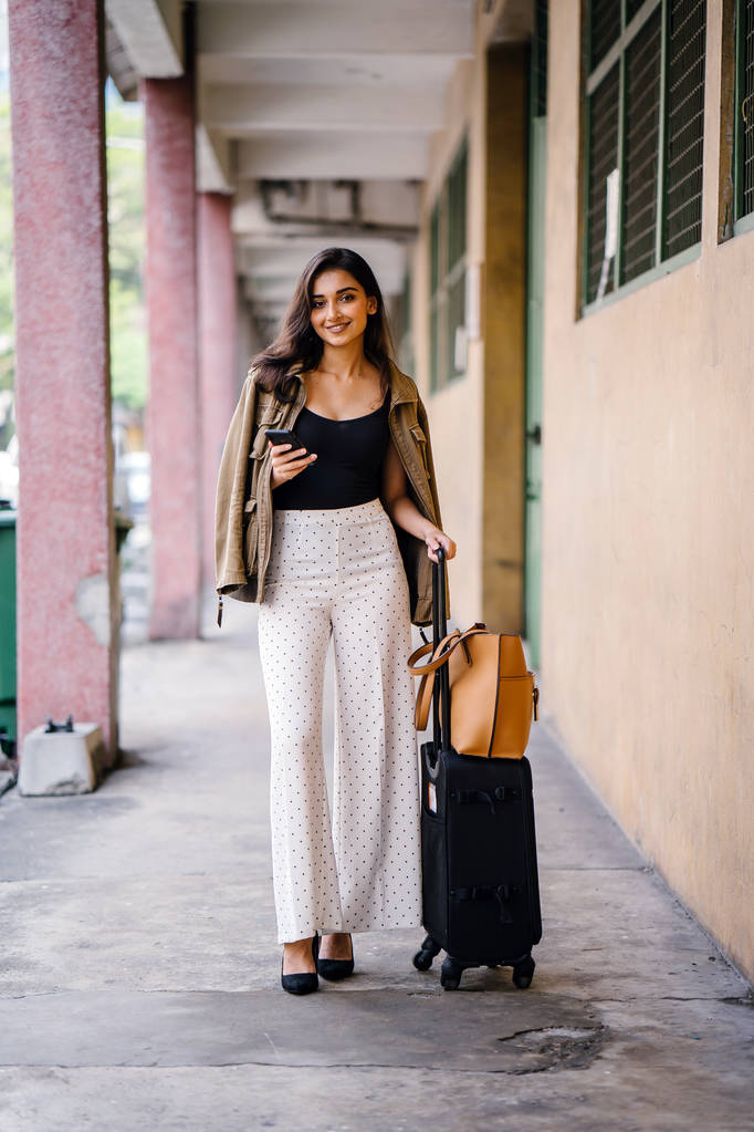 νέοι και ελκυστική ινδική γυναίκα της Ασίας βιβλίων μια βόλτα μέσα από αυτήν ιππεύω χαιρετώντας app στο smartphone της. Εκείνη στέκεται σε μια διάβαση πεζών με τις αποσκευές και κομψά είναι ντυμένος με ενδυμασία και αποχρώσεις - Φωτογραφία, εικόνα