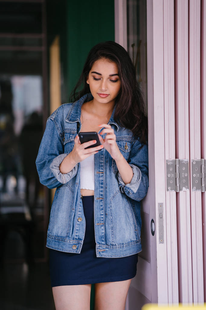 Portrait d'une belle, grande, élégante et jeune femme asiatique indienne textant sur son smartphone. Elle porte une veste en denim rétro
  - Photo, image