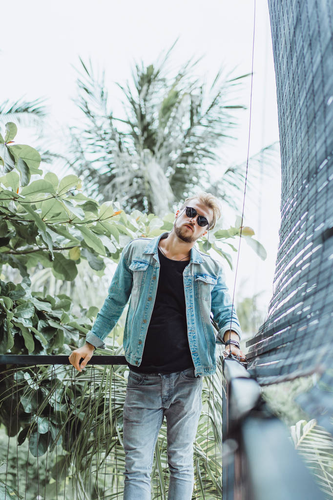nuori houkutteleva mies poseeraa trooppisella paikalla, taustalla palmuja ja vehreyttä, aurinkolasit
 - Valokuva, kuva