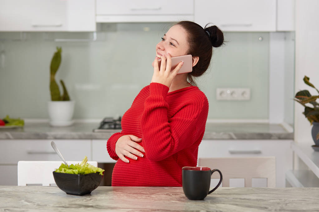 Aufnahme einer attraktiven lächelnden jungen Frau, die sich auf ein Baby freut, die Hand am Bauch behält, per Handy telefoniert, köstlichen frischen Salat isst, lustige Witze hört, am Küchentisch posiert - Foto, Bild