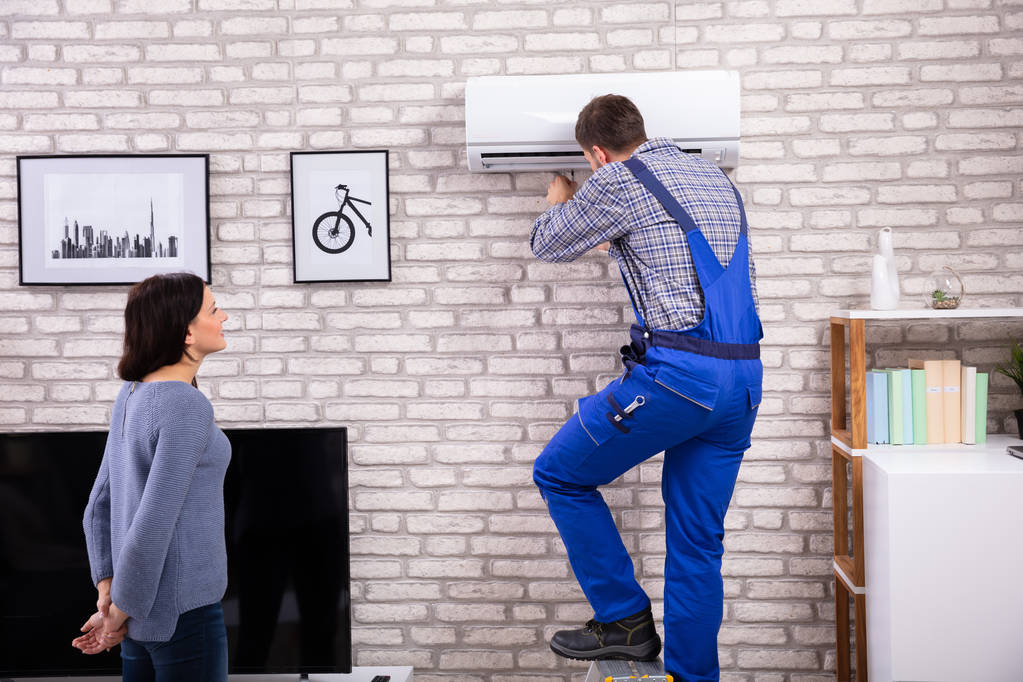 Nuori nainen etsii mies teknikko korjaus ilmastointilaite asennettu tiili seinään
 - Valokuva, kuva
