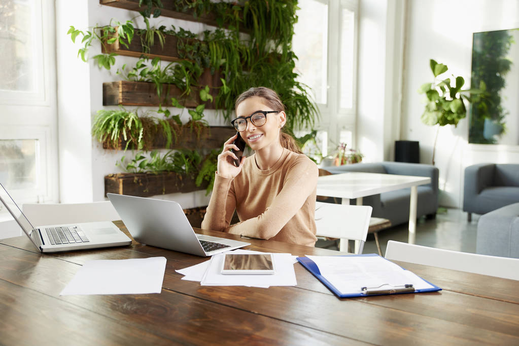 Портрет красивой молодой деловой женщины, сидящей за ноутбуком за столом офиса и разговаривающей с кем-то по мобильному телефону во время работы в офисе
.  - Фото, изображение
