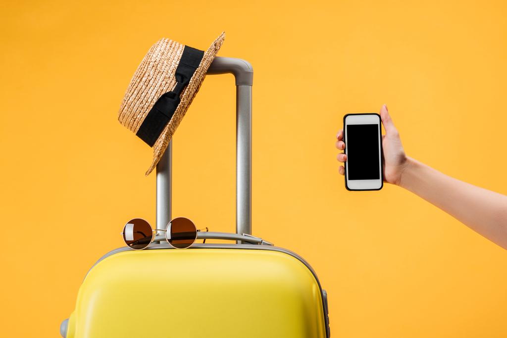cropped view of woman holding smartphone with blank screen near travel bag, straw hat and sunglasses isolated on yellow - Photo, Image