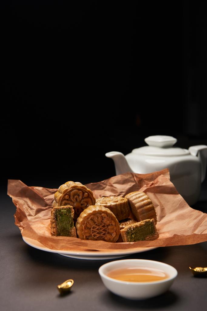 selective focus of traditional chinese mooncakes, tea pot and gold ingots on wooden table isolated on black - Photo, Image