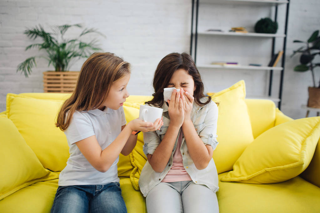 Sick girl on righht sit on sofa and sneeze. She suffer. Girl on left give her white cup. She try to help her. They sit on yellow sofa in room. - Photo, Image