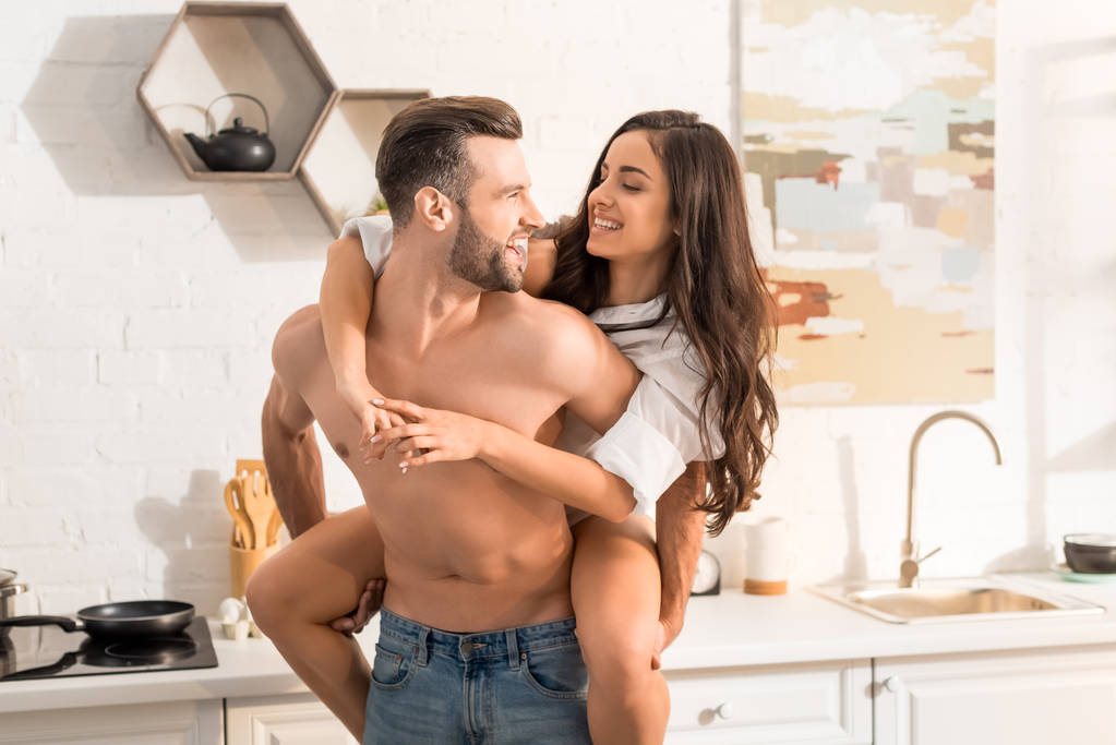 handsome shirtless man giving piggyback ride to smiling woman in kitchen - Photo, Image