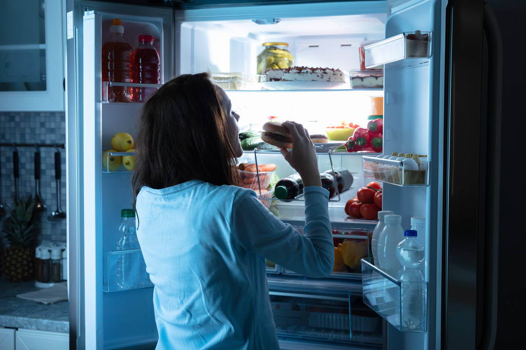 Young Woman Eating Delicious Burger Near Open Refrigerator - Photo, Image