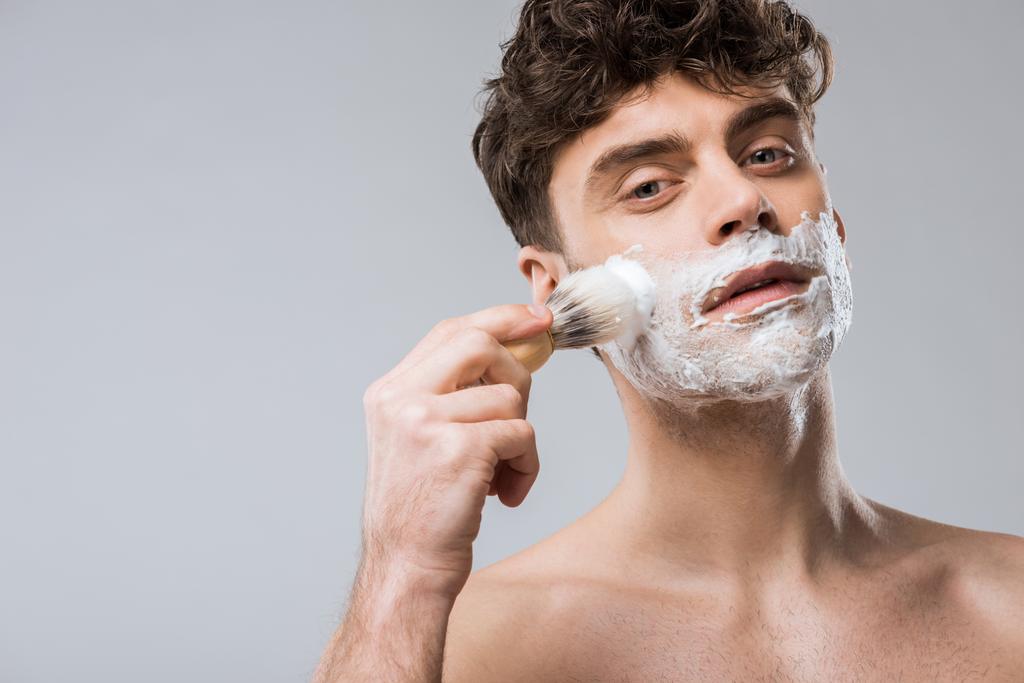 joven guapo aplicando espuma de afeitar con cepillo, aislado en gris
 - Foto, Imagen