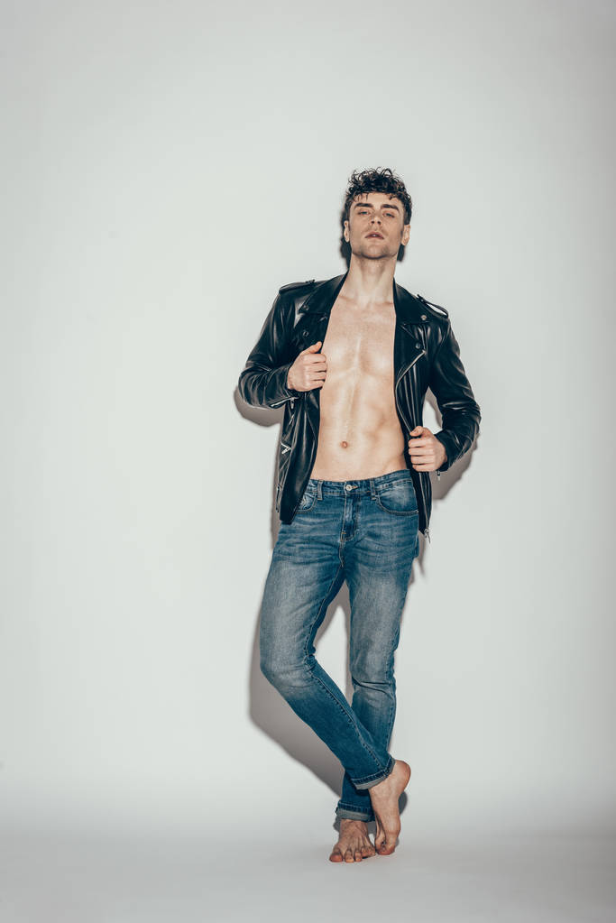 handsome sexy man posing in jeans and black leather jacket on grey - Zdjęcie, obraz