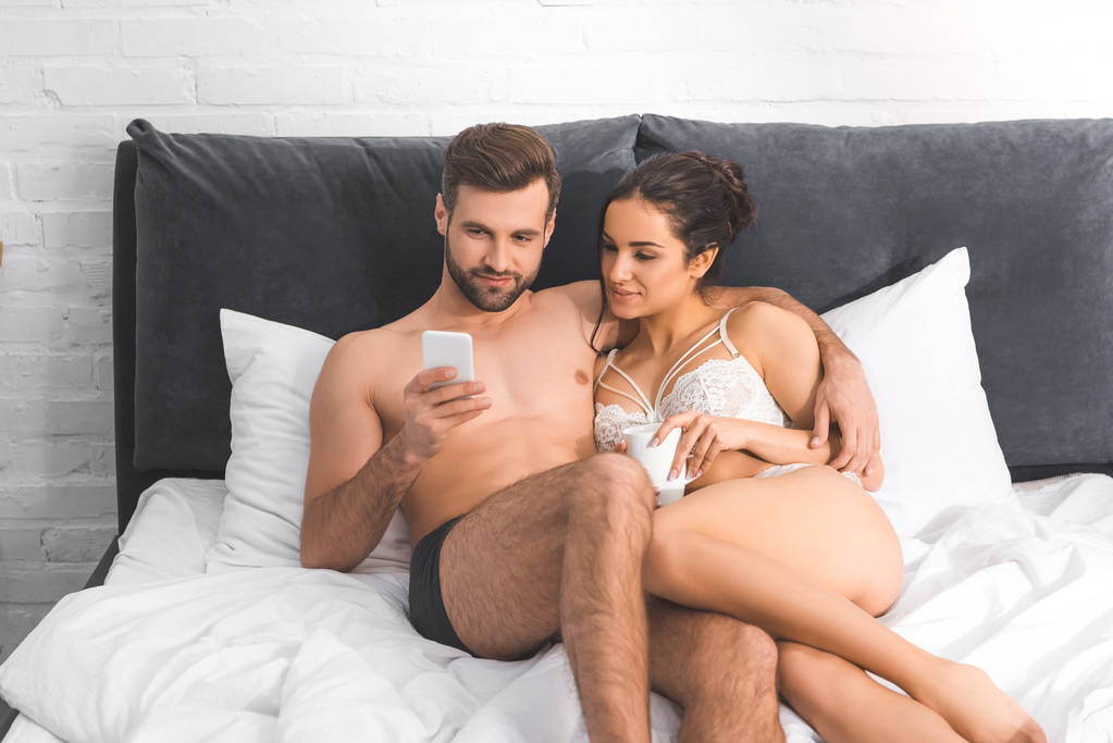 όμορφος άνδρας αγκαλιάζει όμορφη γυναίκα και χρησιμοποιώντας το smartphone στο κρεβάτι - Φωτογραφία, εικόνα