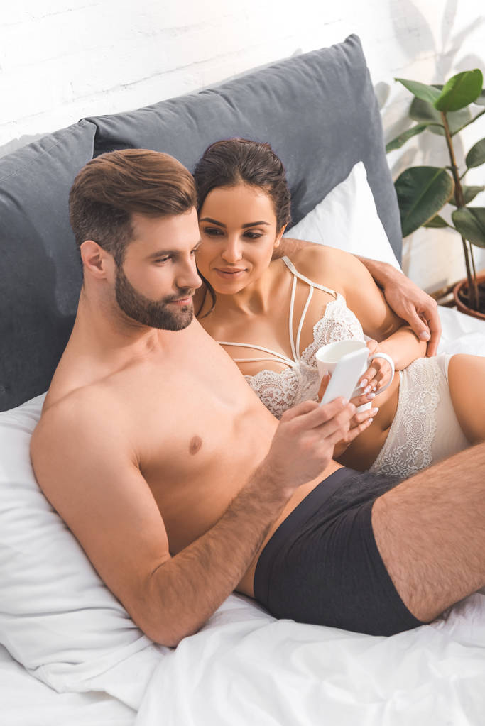 hombre guapo abrazando a mujer hermosa y el uso de teléfono inteligente en la cama
 - Foto, Imagen
