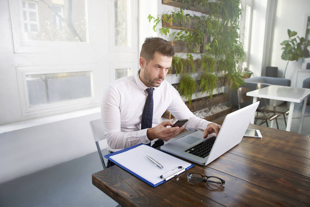 Retrato de hombre de negocios confiado usando camisa y corbata y mensajes de texto mientras está sentado en la oficina y trabajando en el ordenador portátil
. - Foto, imagen
