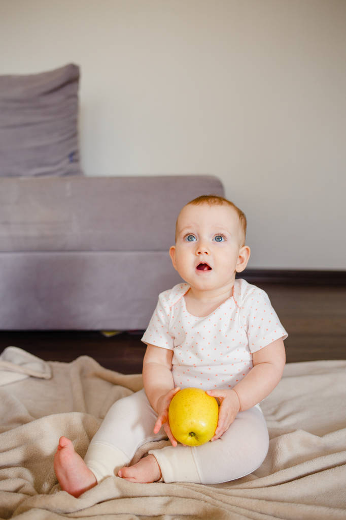Retrato de uma criança pequena bonito com uma maçã amarela, sentado no chão. Bebê menina 9 meses de idade segurando uma fruta
 - Foto, Imagem