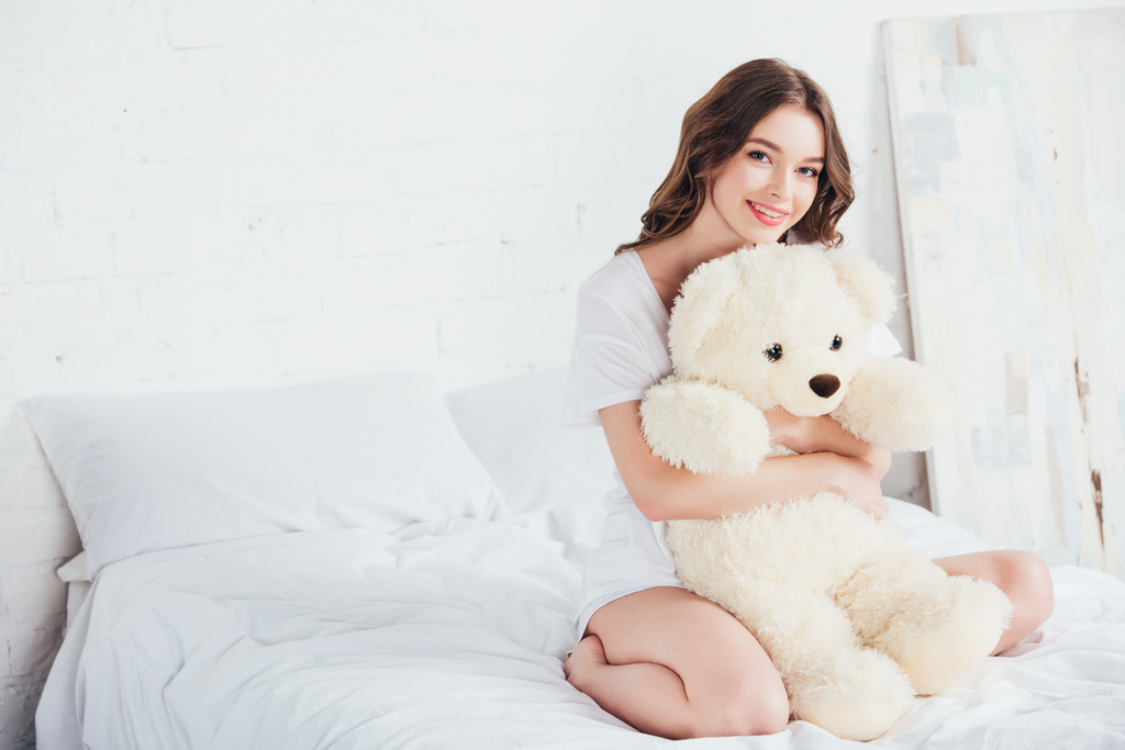 hermosa mujer abrazando osito de peluche y sentado en la cama con ropa de cama blanca
 - Foto, Imagen