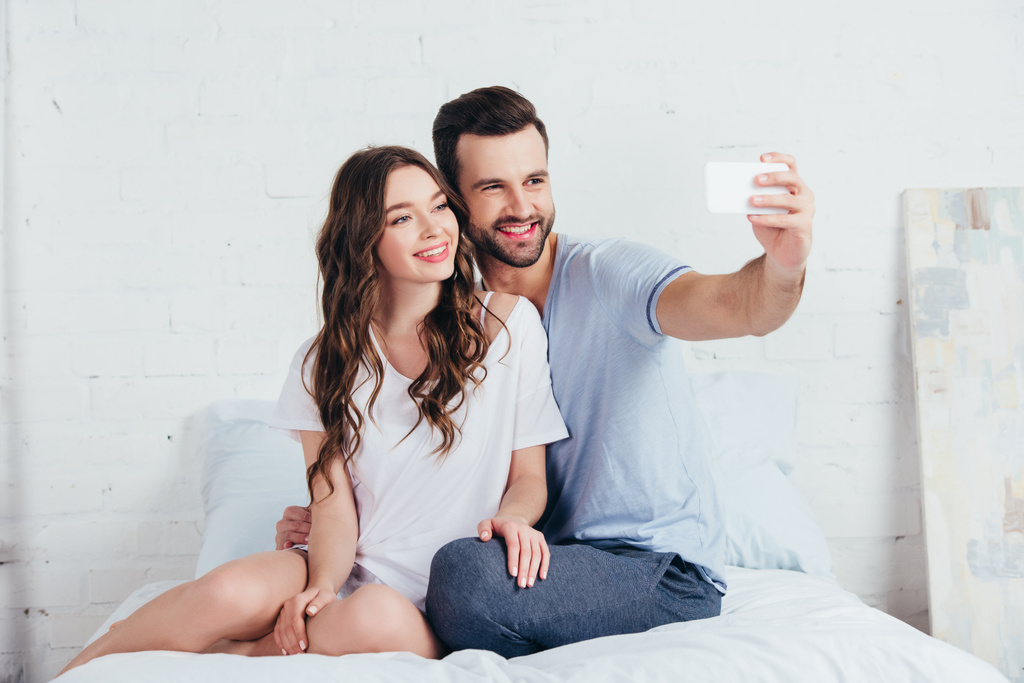 φίλη και φίλο της κάθεται στο κρεβάτι στην κρεβατοκάμαρα και λήψη selfie σε smartphone - Φωτογραφία, εικόνα