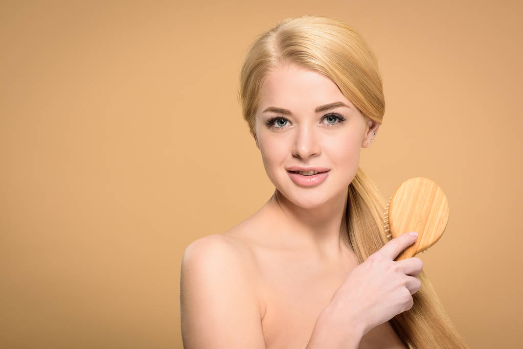 hermosa chica rubia desnuda cepillando el cabello con cepillo de madera y sonriendo a la cámara aislada en beige
  - Foto, imagen