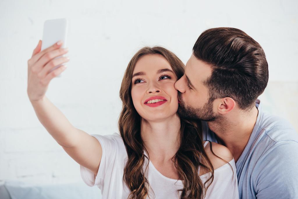 boyfriend kissing girlfriend while taking selfie in bedroom - Photo, Image