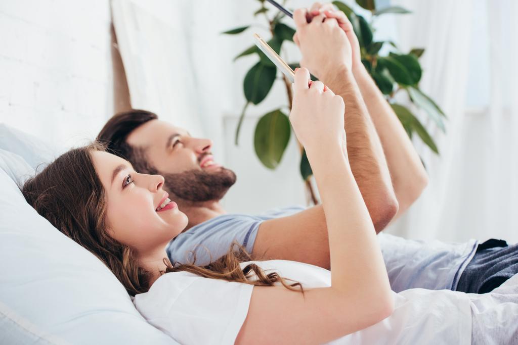jovem casal relaxando na cama enquanto usa smartphones e olhando uns para os outros
 - Foto, Imagem