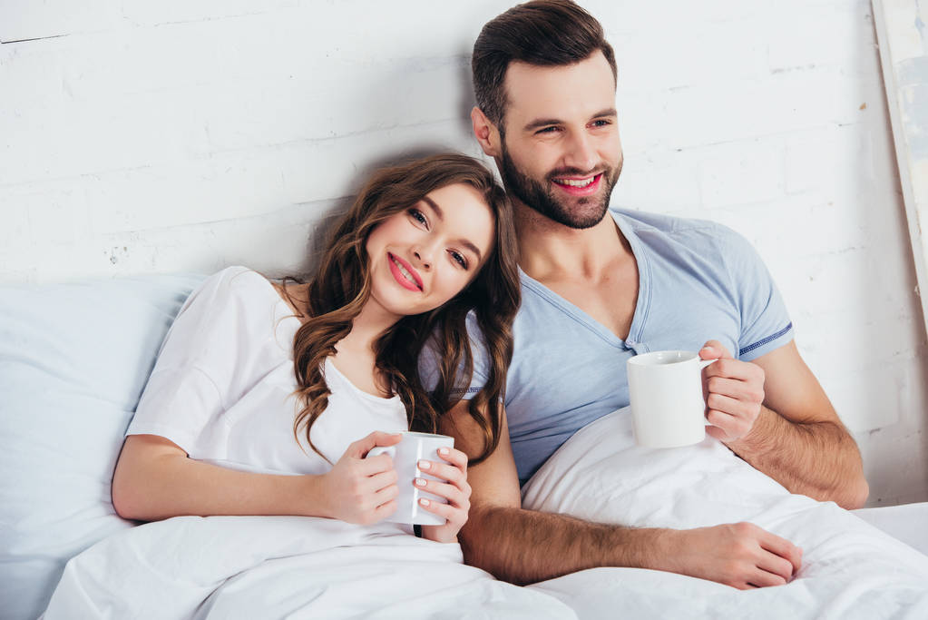 erwachsenes Paar entspannt auf weichem weißen Bettzeug liegend und Tassen haltend - Foto, Bild