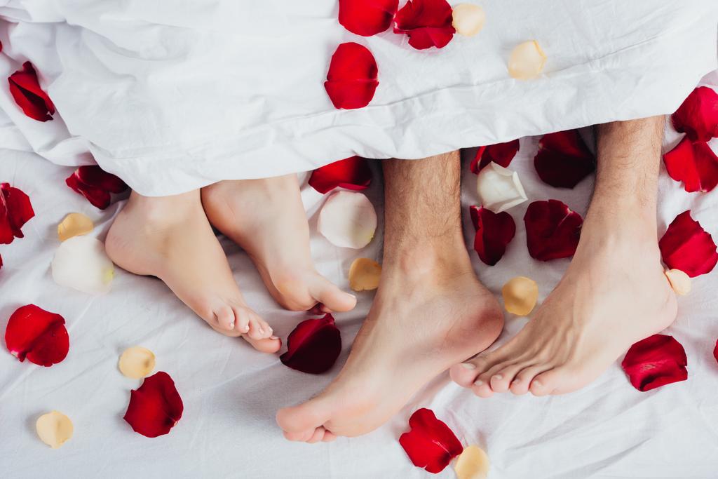 Teilansicht eines barfüßigen Paares, das auf weichem weißen Bettzeug mit roten Blütenblättern liegt  - Foto, Bild