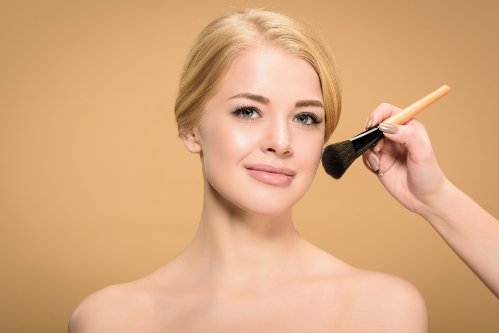 recortado disparo de esteticista aplicación de maquillaje a la joven mujer desnuda mirando a la cámara aislada en beige
  - Foto, imagen