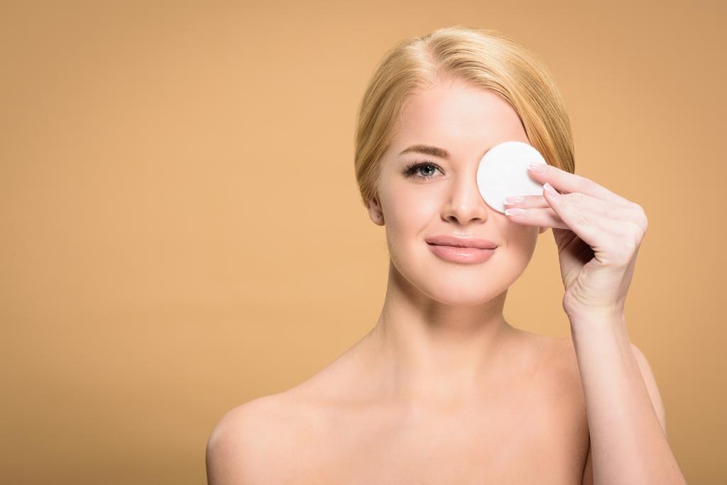 attractive naked woman holding cotton disk near eye and smiling at camera isolated on beige - Photo, Image