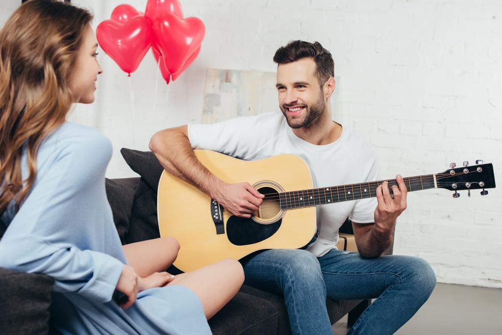 young man playing guitar to smiling girlfriend with heart-shaped balloons on background - Photo, Image