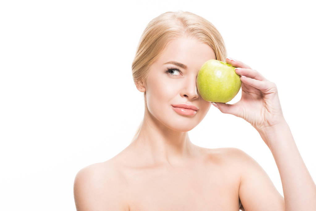 chica rubia desnuda sosteniendo manzana verde cerca del ojo y mirando hacia otro lado aislado en blanco
 - Foto, imagen
