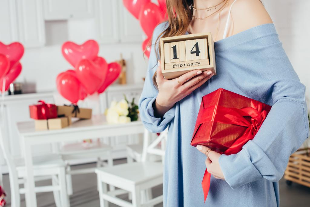 обрізаний вид дівчини, що тримає подарункову коробку та дерев'яний календар з днем святого Валентина вдома
  - Фото, зображення