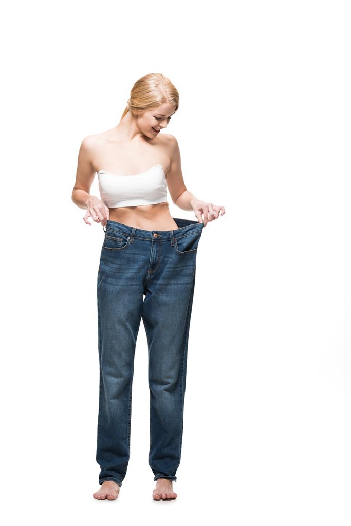 full length view of smiling slim girl wearing oversized jeans and looking down isolated on white - Photo, Image