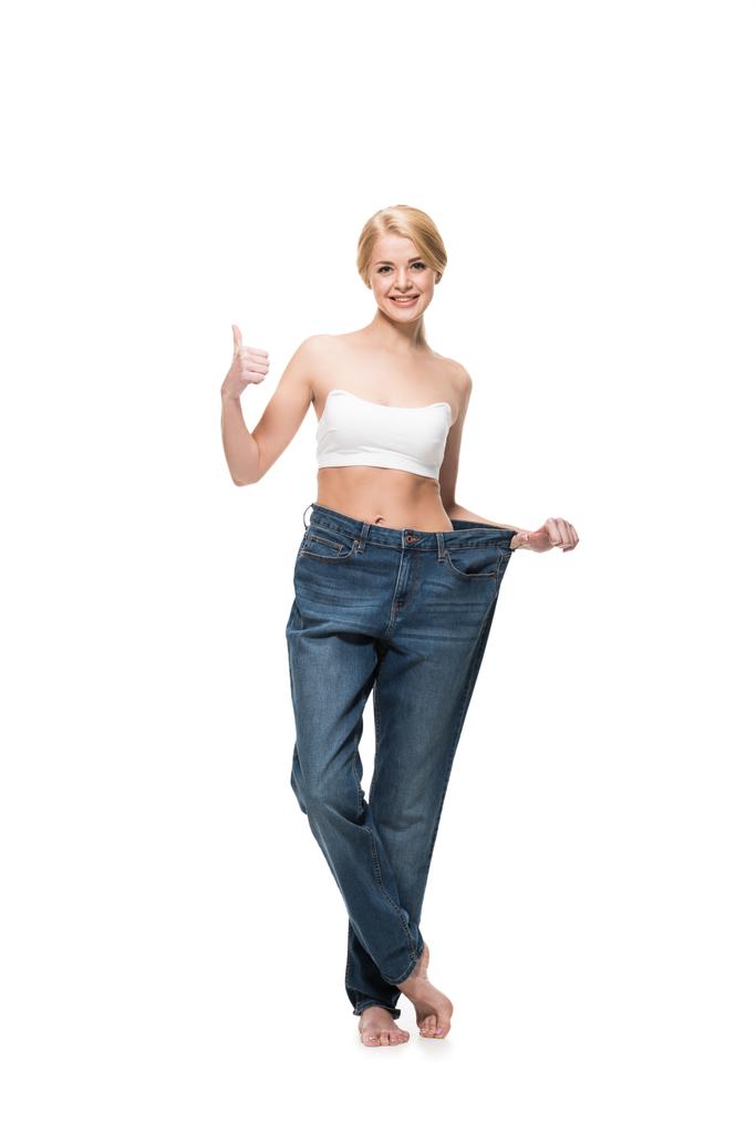 Happy štíhlá žena v nadměrné džíny ukazuje palcem a usmívá se na kameru izolované na bílém - Fotografie, Obrázek