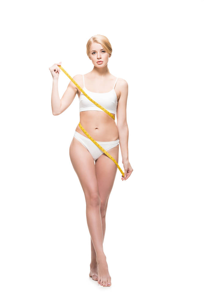 volle lengte weergave van jonge slanke vrouw in ondergoed meetlint rond lichaam en kijken naar camera te houden geïsoleerd op wit - Foto, afbeelding