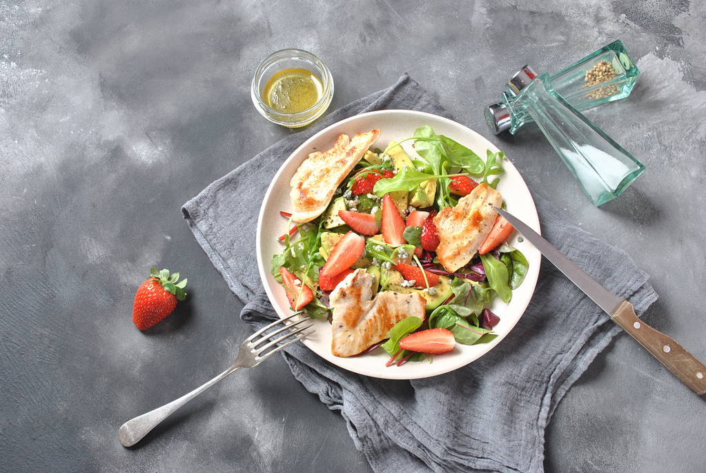 Κοτοσαλάτα με αβοκάντο, μπλε τυρί, φράουλες, ρόκα, φύλλα τεύτλων. Μπολ αβοκάντο υγιεινού γεύματος, πιάτο με κοτόπουλο και φράουλες για το σκοτεινό γκρι φόντο. Έννοια της υγιεινής διατροφής - Φωτογραφία, εικόνα