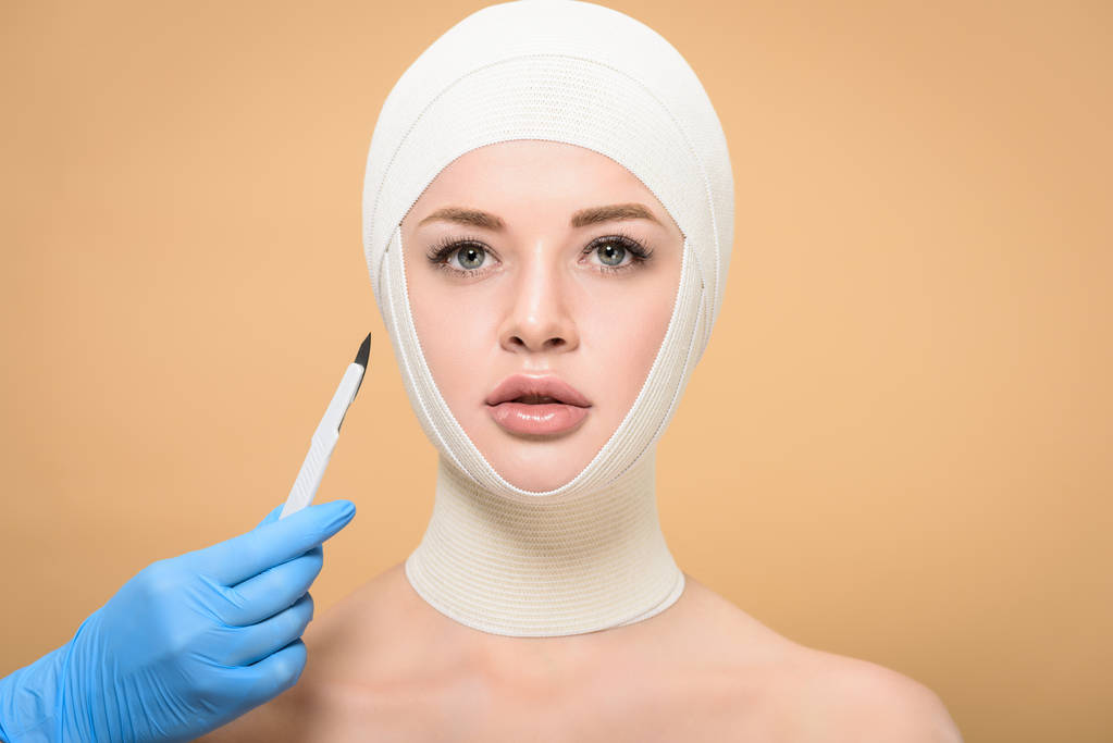 inyección recortada de médico sosteniendo el bisturí cerca de la mujer con vendajes sobre la cabeza aislada en beige
 - Foto, Imagen