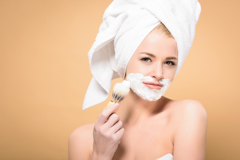 young woman with towel on head applying foam with shaving brush isolated on beige - Photo, Image