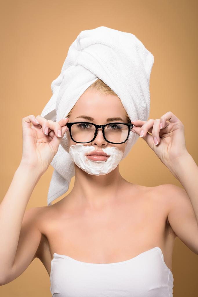 woman with towel on head and shaving cream on face adjusting eyeglasses and looking at camera isolated on beige - Photo, Image