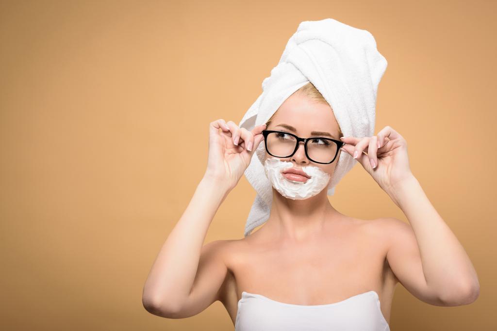junge Frau mit Handtuch auf dem Kopf und Rasierschaum auf der Gesichtsbrille und isoliertem Blick auf beige - Foto, Bild