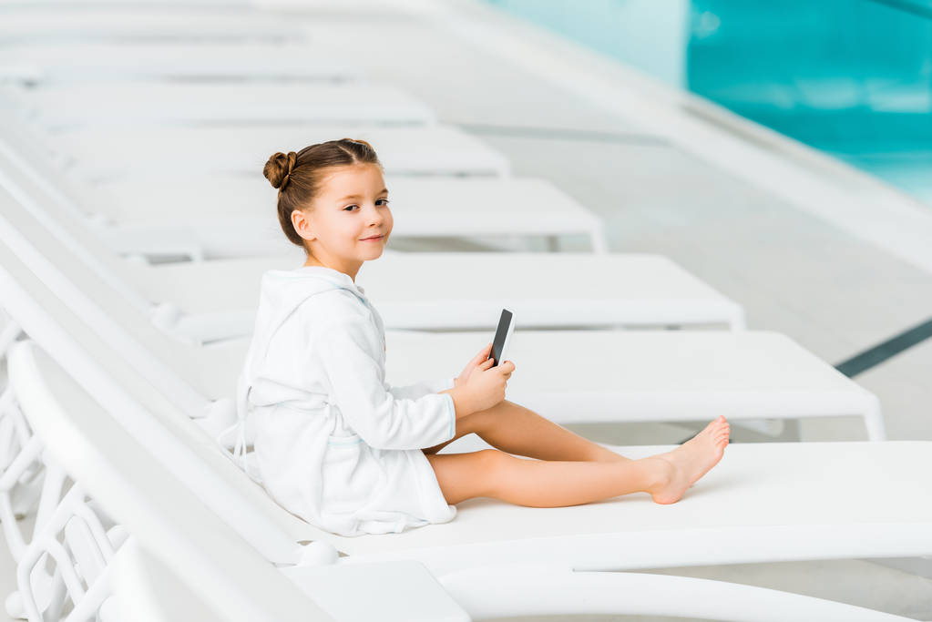 милый ребенок в белом халате с смартфоном возле бассейна
 - Фото, изображение